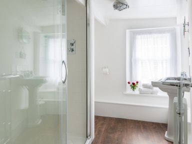 A bathroom with a glass shower enclosure a window with sheer curtains a pedestal sink and wooden flooring at Bre Cottage in Port Isaac
