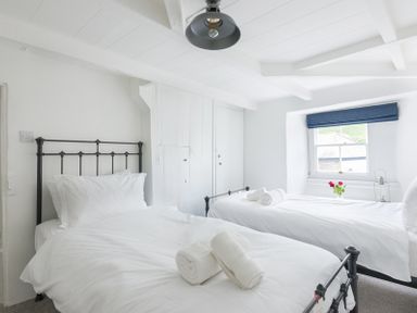 A bedroom with two single beds with white bedding and rolled towels a window with a blue blind a ceiling light and built in cupboards at Bre Cottage in Port Isaac
