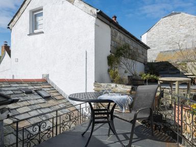 A small outdoor balcony with a round metal table and one chair overlooking stone roofs and white buildings at Bre Cottage in Port Isaac