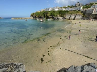 A beach with people walking and playing near clear water with boats and houses on a cliff at Bre Cottage in Port Isaac