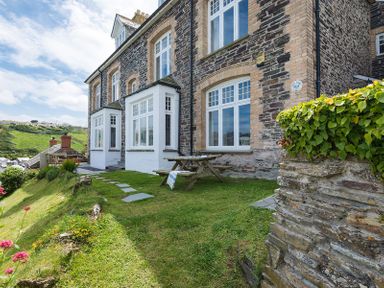 An outdoor area with a picnic table on grass next to a stone house with multiple windows and a stone wall with greenery at Folly 2 in Port Isaac
