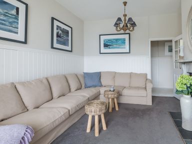 A living room with a beige sectional sofa two small wicker stools framed ocean pictures on the walls and a chandelier at Folly 2 in Port Isaac