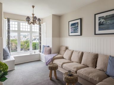 A living room with a beige sofa blue and striped cushions wicker stools framed ocean pictures and bay window seating at Folly 2 in Port Isaac