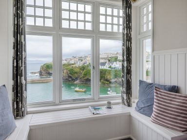 A window seat with cushions and a book with a mug on the ledge overlooking water with boats and houses at Folly 2 in Port Isaac
