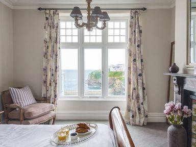 A bedroom with a bed holding breakfast tray next to a window with floral curtains and a chair at Folly 2 in Port Isaac