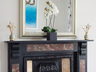 A black fireplace with decorative tiles and a large framed mirror above with flowers on the mantel at Folly 2 Port Isaac