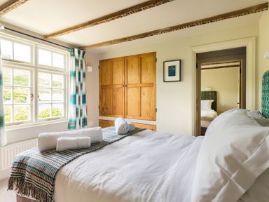 A bedroom with a made bed holding rolled towels beside a window with striped curtains and wooden built-in wardrobes at Crow's Nest in Port Isaac