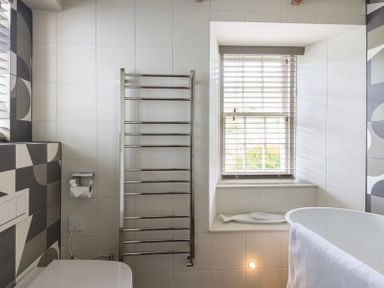 A bathroom with a white toilet a metal towel rack a window with blinds and a white bathtub with a towel at Crows Nest in Port Isaac