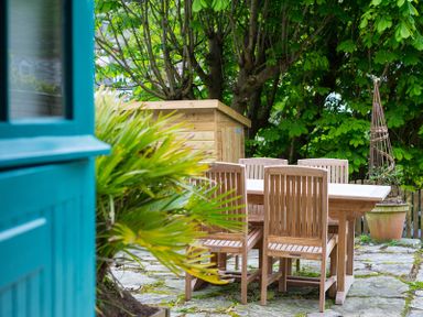 An outdoor patio with a wooden dining table and chairs surrounded by green plants and a wooden shed in the background at Crow's Nest in Port Isaac