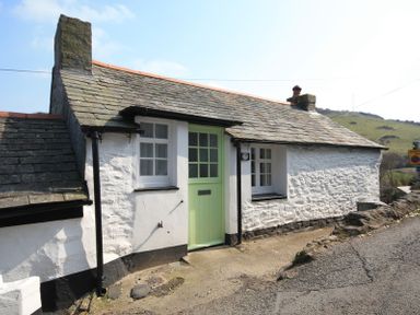 A stone cottage with a green door and two windows beside a road with grassy hills in the background at Crow's Nest in Port Isaac
