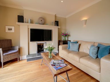 A living room with a beige sofa and blue pillows a wooden coffee table with magazines and flowers a fireplace with a mounted TV and a wicker chair at Worthy House in Daymer Bay