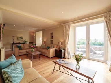 A living room with beige sofas wooden tables flowers and glass doors leading to an outdoor area at Worthy House in Daymer Bay