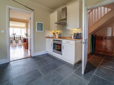 A kitchen with white cabinets and an oven next to a room with a dining table and chairs at Worthy House in Daymer Bay