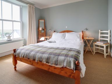 A bedroom with a wooden bed with white bedding a plaid blanket bedside tables with lamps a chair a standing mirror and a window at Worthy House in Daymer Bay