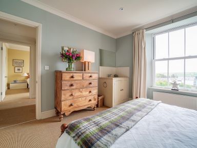 A bedroom with a bed, wooden chest of drawers with a lamp and flowers, a small sink, and a window with curtains at Worthy House in Daymer Bay