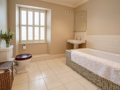 A bathroom with a bathtub with a white towel a sink with a mirror a toilet with a wicker basket and a window with shutters at Worthy House in Daymer Bay