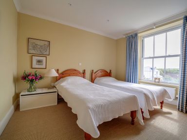 A bedroom with two single beds white bedspreads bedside table with lamp and flowers window with check curtains at Worthy House in Daymer Bay