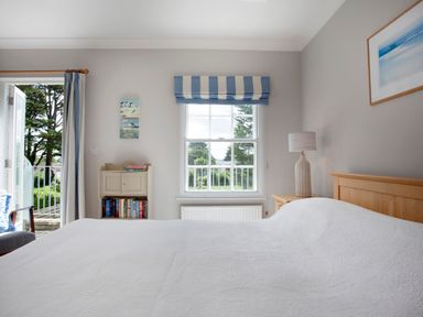 A bedroom with a bed covered by a white bedspread a wooden nightstand with a lamp a window with a blue and white striped valance a small cabinet with books and paintings on the wall at Lowenna Manor 4 in Rock