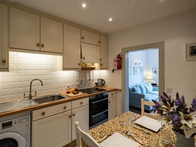 A kitchen with white cabinets wooden countertops a stove and a sink next to a dining table with a floral tablecloth and flowers at April Cottage in Port Isaac