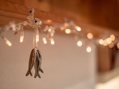 Two metallic fish ornaments hanging from a string attached to wooden beam with string lights in the background at April Cottage in Port Isaac