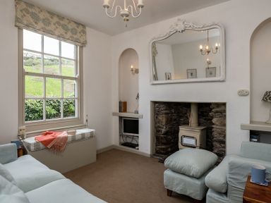 A sitting room with a window seat a wood burner in a stone fireplace and a large mirror above at April Cottage in Port Isaac