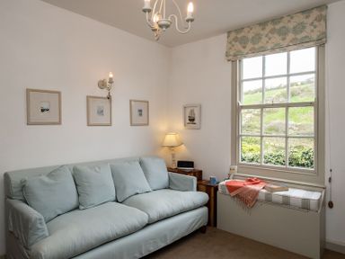 A room with a light blue sofa a window seat with a cushion and throw a lamp and framed pictures on the wall at April Cottage in Port Isaac