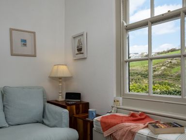 A corner of a room with a light blue armchair a lamp on a wooden side table a large window and a cushioned bench with a book and an orange throw at April Cottage in Port Isaac