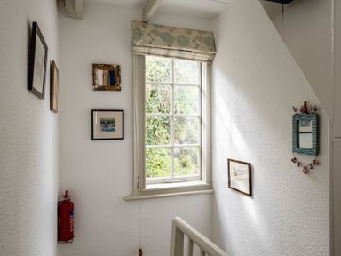 A stairwell with a window showing outside greenery and framed pictures on the walls at April Cottage in Port Isaac