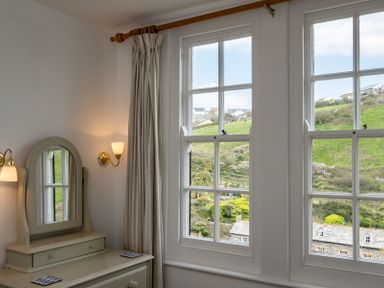 A room with a dresser with mirror next to windows showing a hillside at April Cottage in Port Isaac