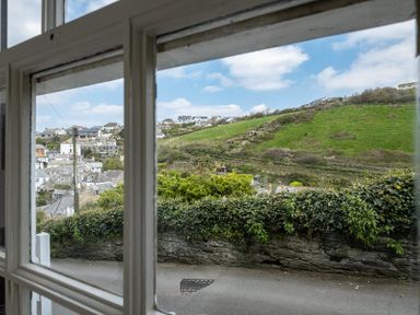 A window view overlooking a road with a stone wall and greenery with houses and hills in the background at April Cottage Port Isaac