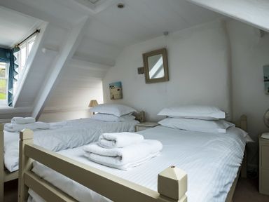 A bedroom with two single beds white linens white towels on the beds a window with blue curtains a bedside table and a mirror on the wall at April Cottage in Port Isaac