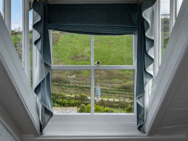 A window with blue curtains and a small hanging decoration showing a green hillside at April Cottage in Port Isaac
