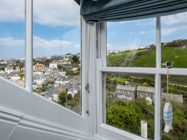 A view of houses and green hills seen through a window with white frames and a blue striped fish decoration at April Cottage in Port Isaac