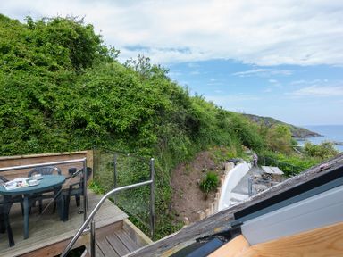 An outdoor wooden deck with a table and chairs next to a hillside overlooking the sea at April Cottage in Port Isaac