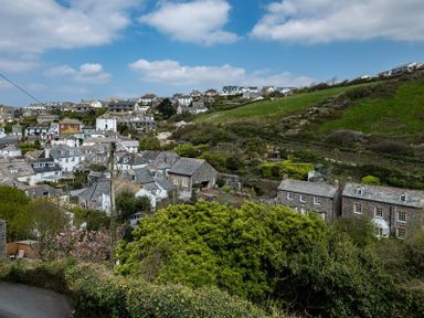 A hillside village with stone houses surrounded by greenery and a partly cloudy sky at April Cottage in Port Isaac