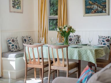 A dining area with a table covered with a green polka dot tablecloth set with plates and glasses surrounded by wooden chairs and a bench with fish patterned cushions at Carn Du 1 in Rock