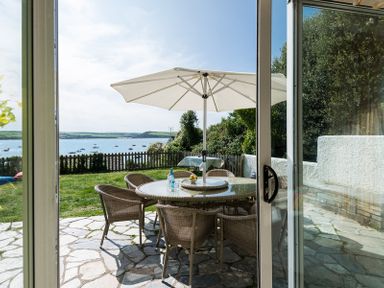 A patio with a round glass table with six wicker chairs and a large white umbrella near a grassy lawn and water at Carn Du 1 in Rock