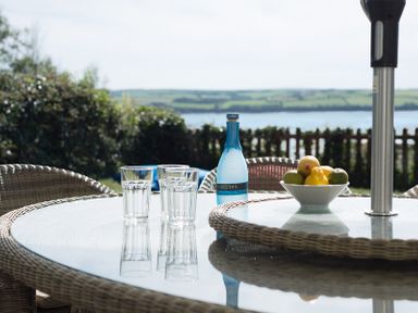 A glass outdoor table with three drinking glasses a blue bottle a bowl of fruit and wicker chairs in a garden with a water view at Carn Du 1 in Rock