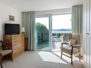 A room with a wooden dresser holding a television beside a wooden chair and an armchair with a cushion next to sliding glass doors with a view of the sea at Carn Du 1 in Rock