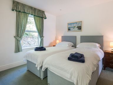 A bedroom with two single beds with folded towels on each bed two bedside tables with lamps a window with green checked curtains and a painting on the wall at Carn Du 1 in Rock