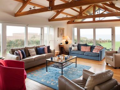 A living room with wooden beams sofas armchairs a coffee table and large windows overlooking greenery at Ossco Polzeath