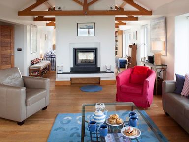A living room with a fireplace four blue mugs a red armchair and a glass coffee table at Ossco in Polzeath