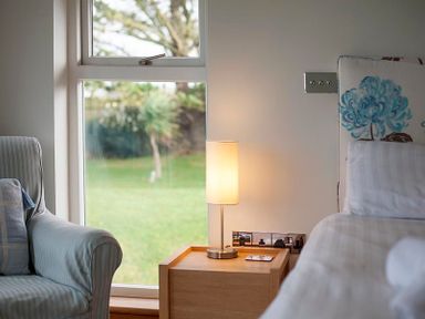 A bedside table with a lamp next to a bed and an armchair by a window at Ossco in Polzeath