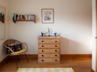 A room with a wooden chest of drawers with sculptures on top a wicker chair with cushions a wall shelf with books and a framed picture on the wall at Ossco in Polzeath