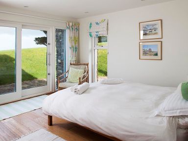 A bedroom with a bed white linens a wicker chair and glass doors opening to a garden at Ossco in Polzeath