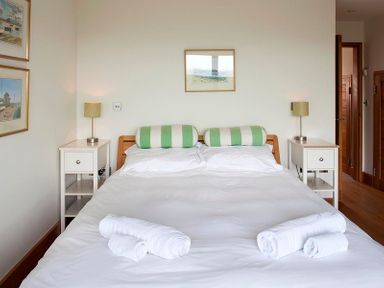 A bedroom with a double bed with white bedding and green striped pillows two white bedside tables with lamps and framed pictures on the walls at Ossco in Polzeath