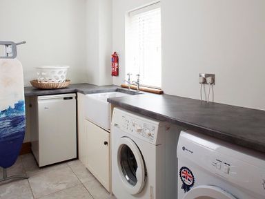 A utility room with washing machine dryer sink ironing board and laundry basket at Ossco in Polzeath