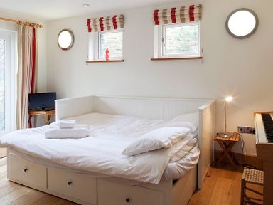 A bedroom with a white daybed with pillows and folded towels next to a wooden side table with a lamp and a small TV on a stand at Ossco in Polzeath