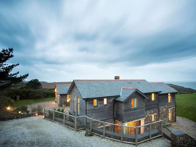 A two-story house with mixed wood and stone exterior and lit windows surrounded by grass and trees at Ossco in Polzeath