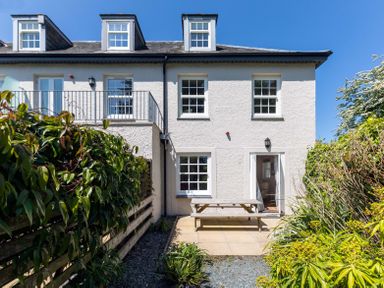 The back of a white two-story house with three dormer windows a patio door and windows a wooden picnic table on a paved patio and a garden with plants and shrubs at Lowenna Manor 10 Rock